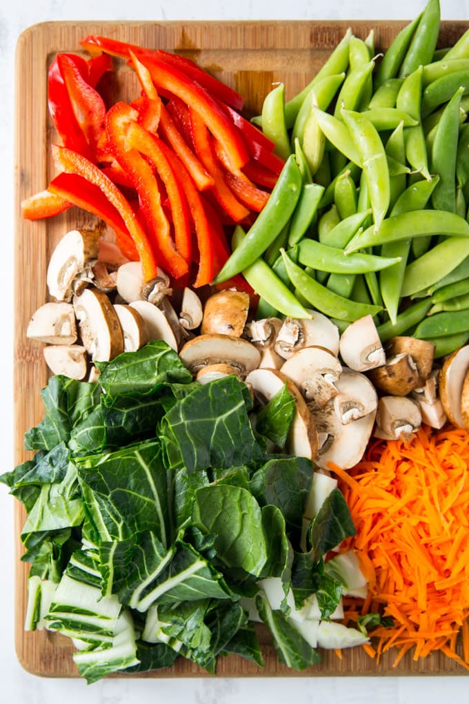 Chopped up bell pepper, mushrooms, bok choy, carrots and snow peas on a wooden cutting board