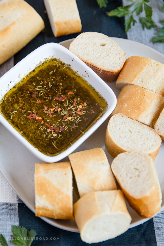 Serve this spicy Garlic & Herb Olive Oil Bread Dip with a crusty french baguette or crostini and you will be the talk of the dinner table. #appetizer a plate of sliced french baguette with a bowl of olive oil bread dip