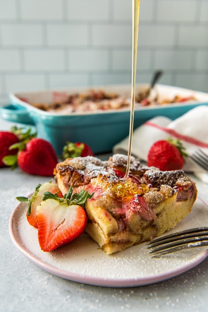 A piece of baked french toast with strawberries and a strem of syprup being poured on top.