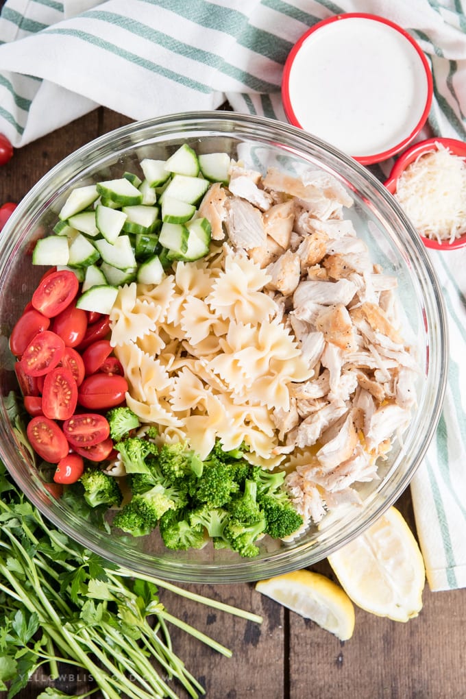 large glass bowl, with chicken, tomatoes, broccoli, cucumbers, pasta, green and white napkin, parsley, small red bowls with white dressing