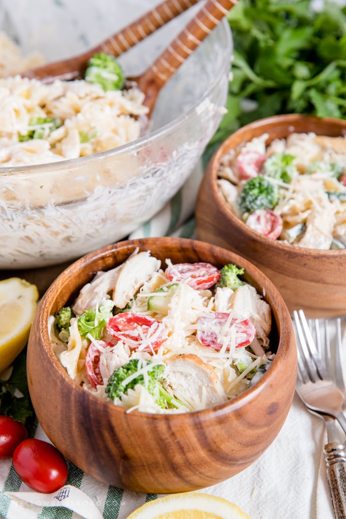 45 degree angle shot of chicken pasta salad in wood bowls and in a large glass bowl. wooden spoons parsley