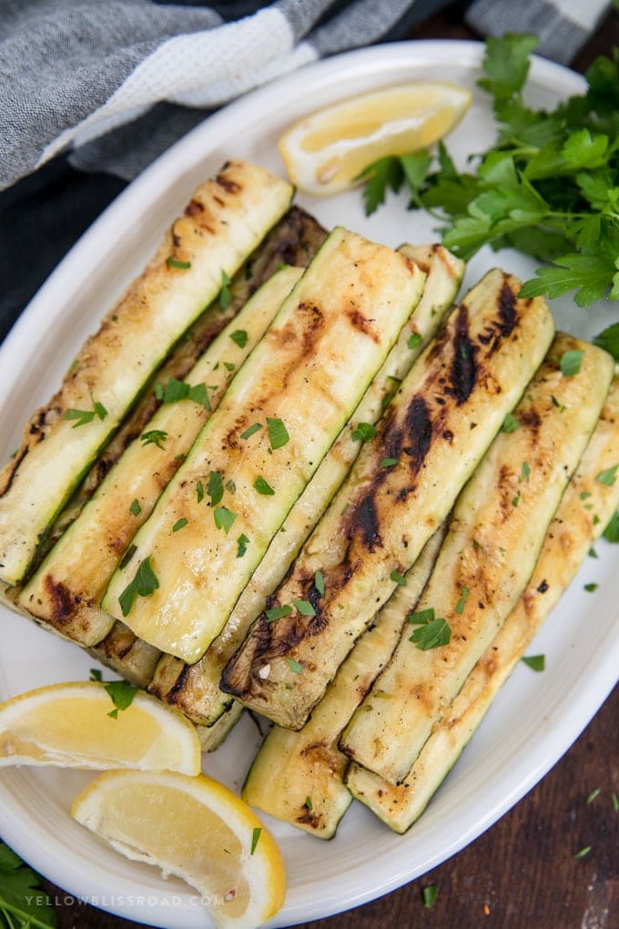 Grilled Zucchini, marinated in lemon juice and olive oil, is a healthy summer side dish that everyone loves! Zucchini with grill marks cut into strips on a plate with lemon and parsley.