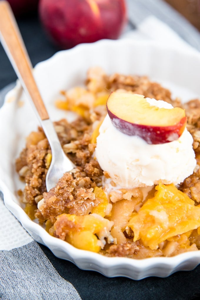 A serving of fresh peach crisp in a bowl with a fork and a scoop of vanilla ice cream