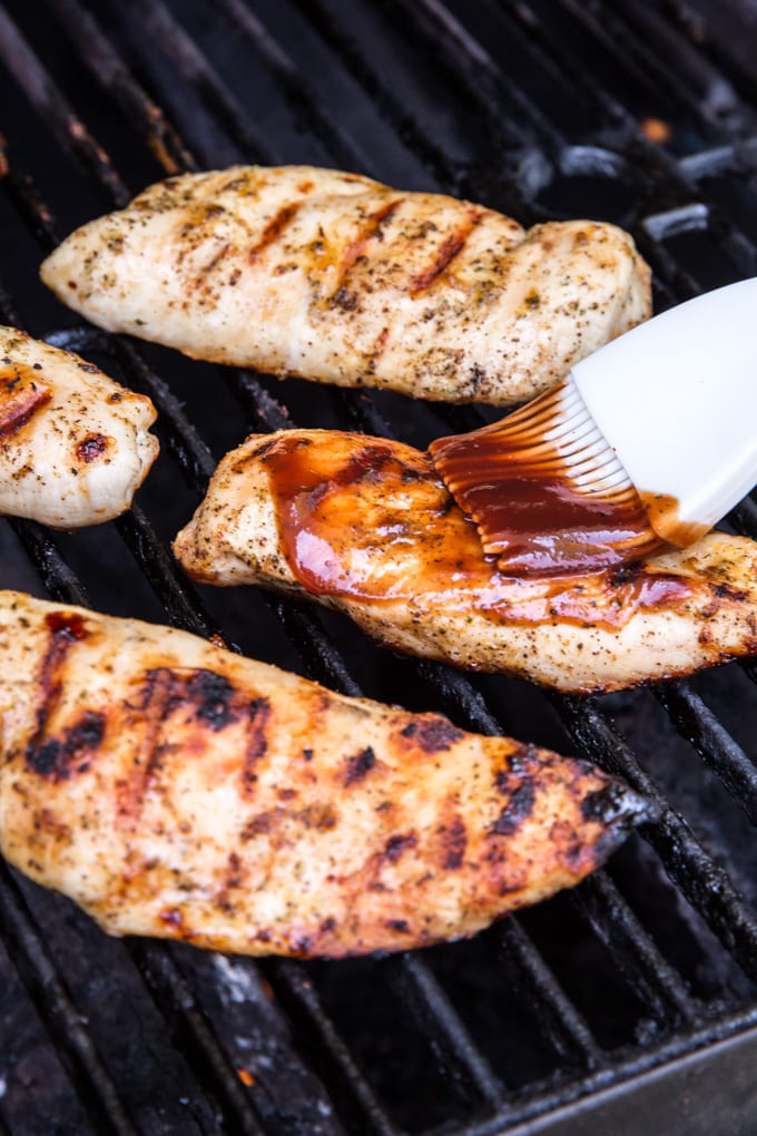 Black grill grates, 4 chicken breasts with grill marks, a white pastry brush with bbq sauce