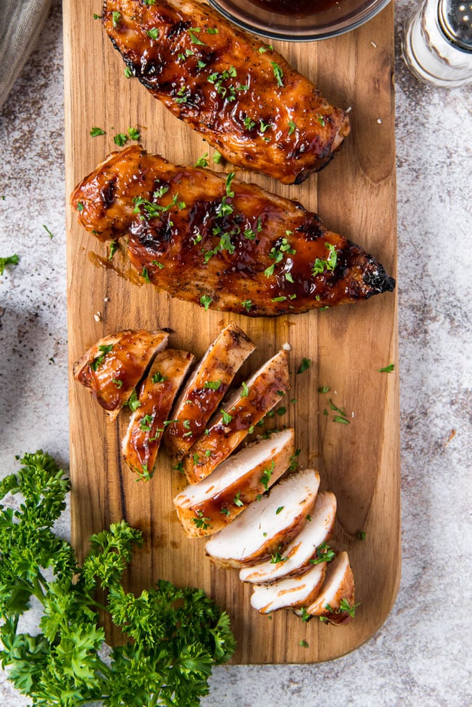 a wooden cutting board, 2 chicken breasts with bbq sauce and parsley, 1 chicken breast, sliced with bbq sauce and parsley, a sprig of parsley