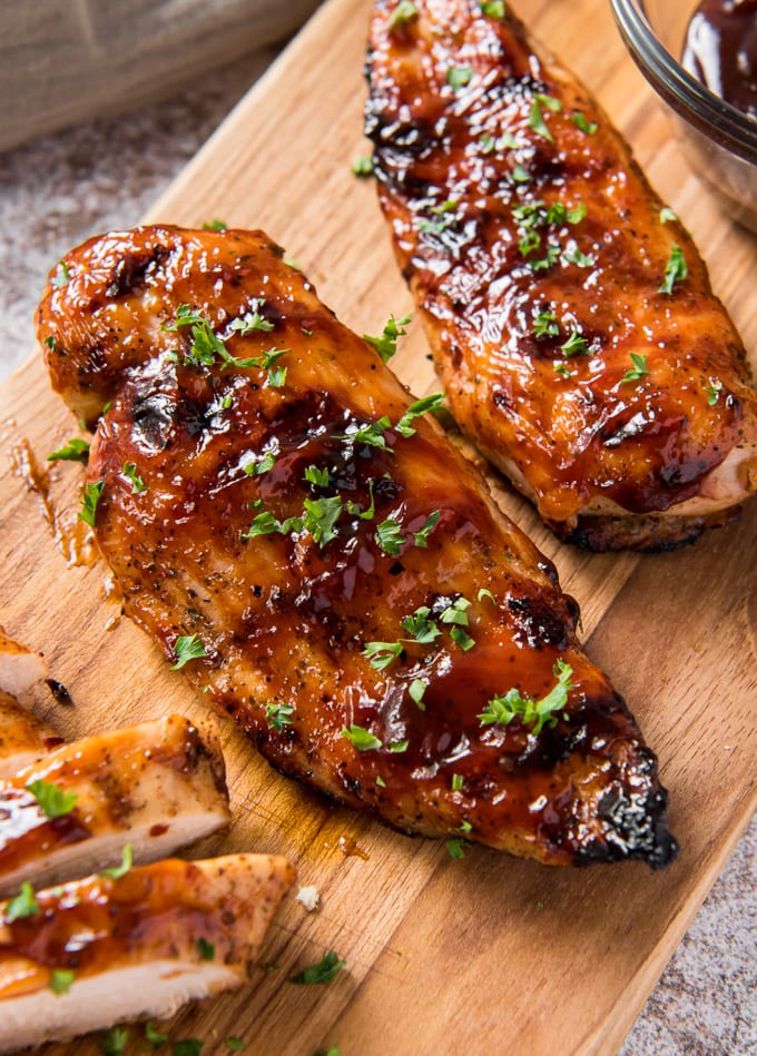 2 chicken breast with bbq sauce and parsley, a couple pieces of sliced chicken, a wood cutting board