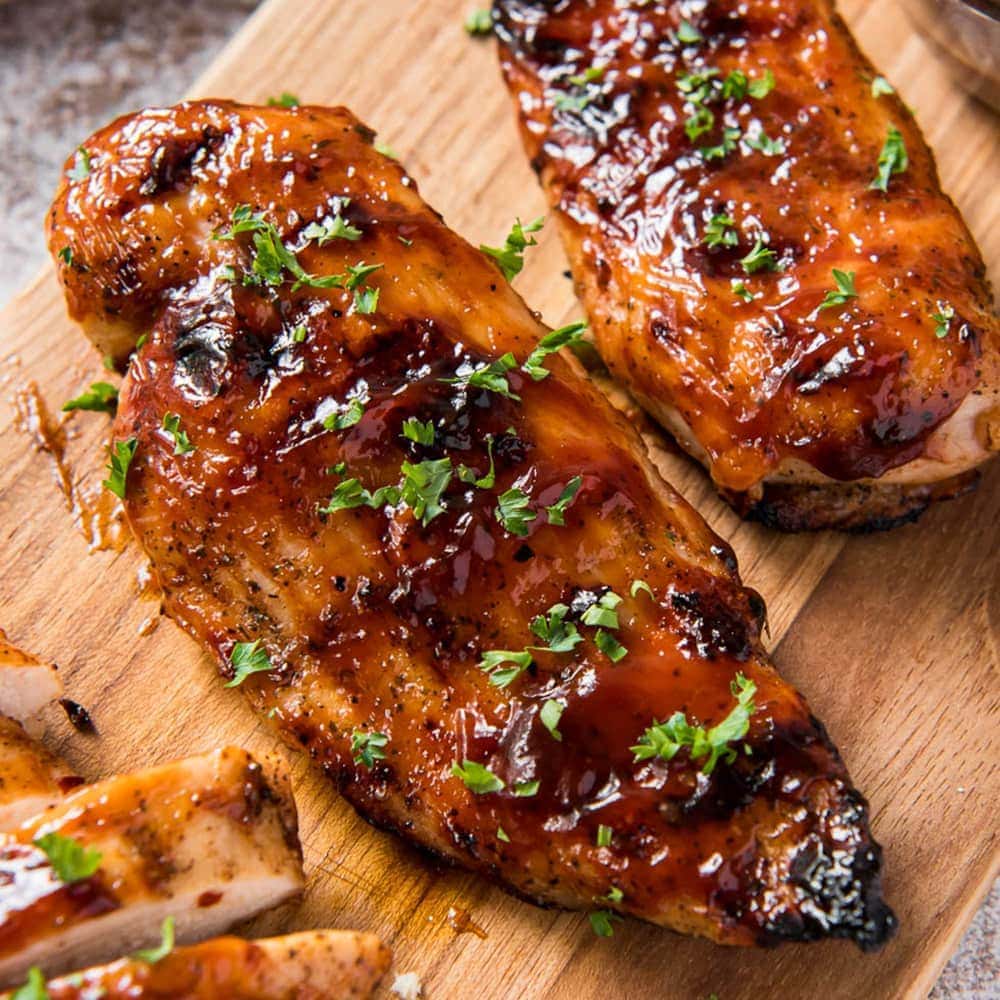 Two grilled chicken breasts covered in barbecue sauce on a wooden cutting board.