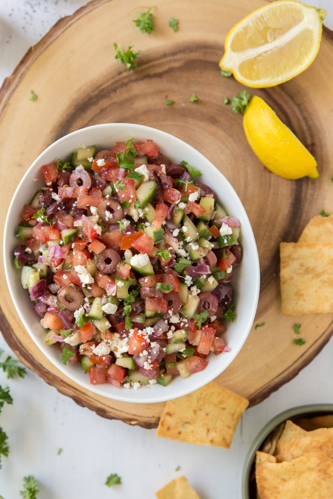 a large, round wood board with a small bowl of greek salsa, surrounded by a few pita chips and lemon slices.