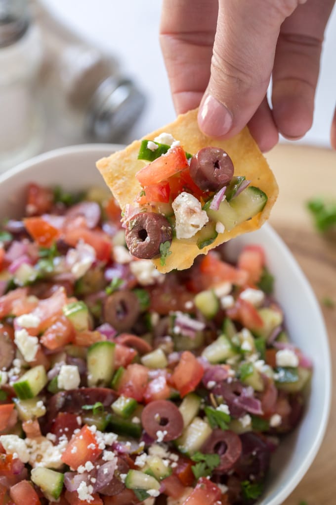 a hand holding a pit chip with diced cucumber, tomatoes, olives and feta cheese. Below is a bowl filled with a larger portion of the same.