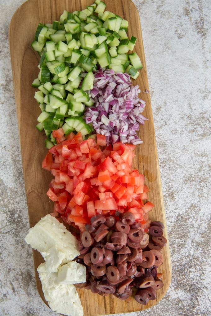 a rectangular shaped wooden cutting board with chopped cucumbers, onion, tomatoes, olives and feta cheese on a gray and white background