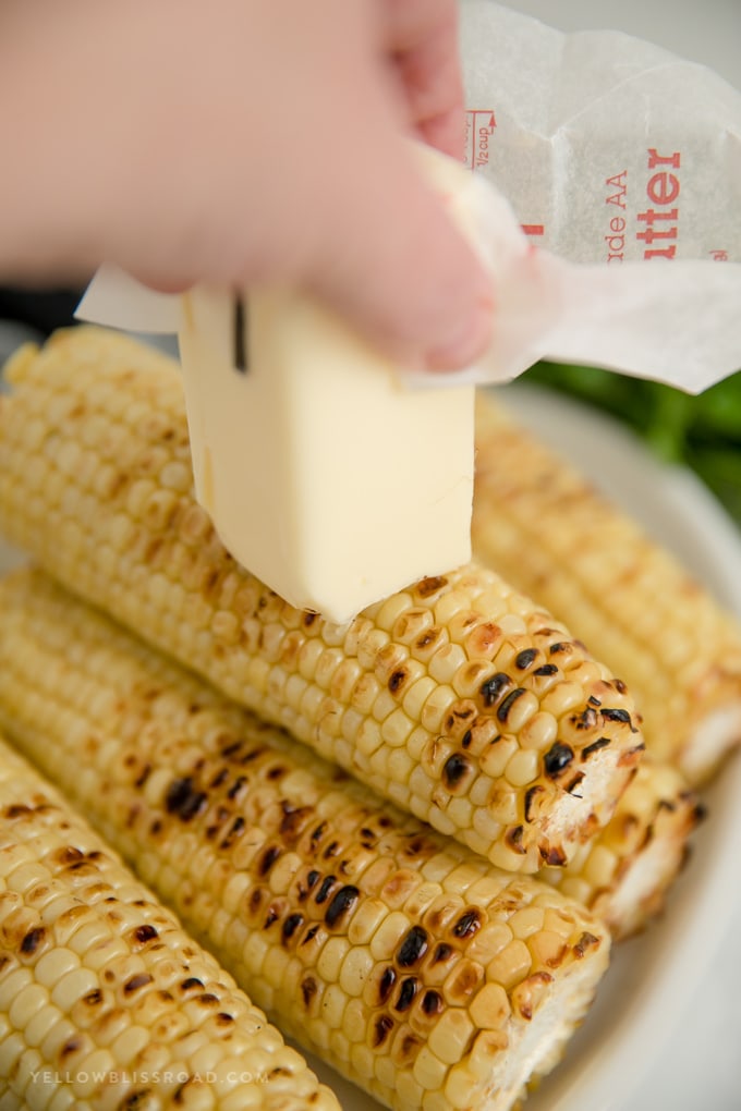 A hand holding a stick of butter rubbing it onto a grilled ear of corn
