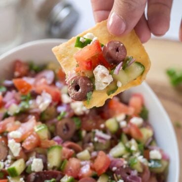 A person holding a cracker with mixture of feta, kalamata olives, tomatoes, and cucumbers.