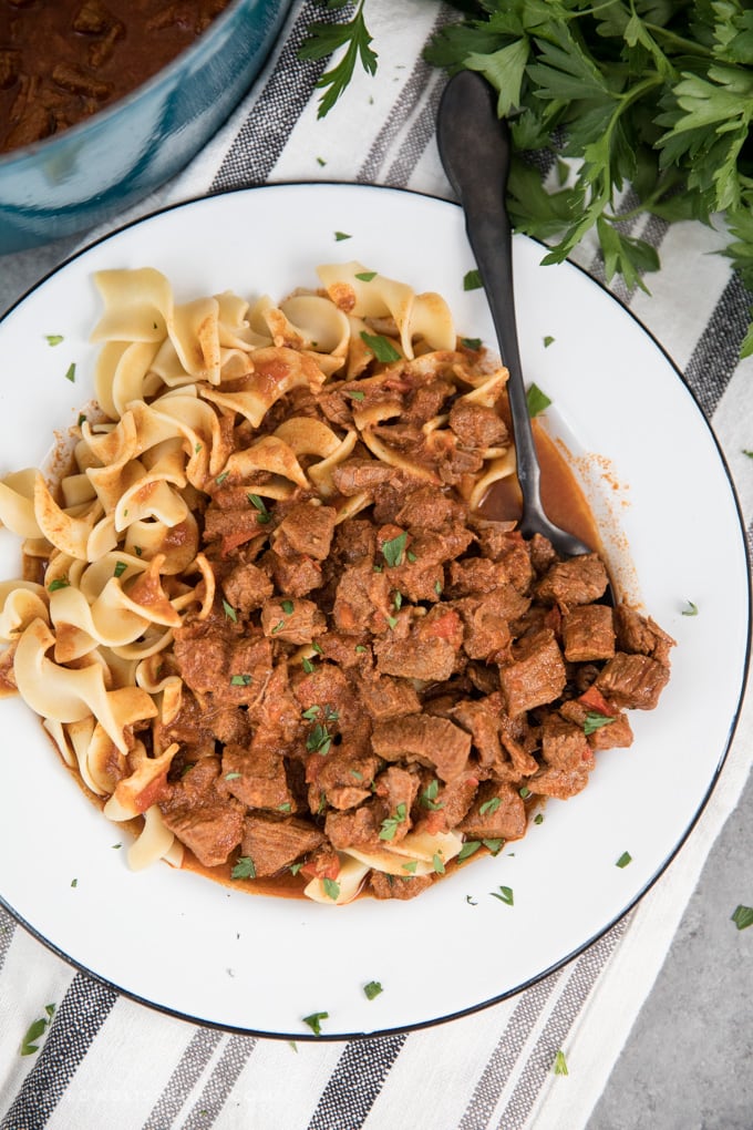 This Classic Hungarian Goulash recipe is a delicious dinner that's packed full of slow-cooked flavor with tender chunks of beef in a rich, thick broth. Authentic Hungarian Goulash in a shallow white bowl with a black spoon.