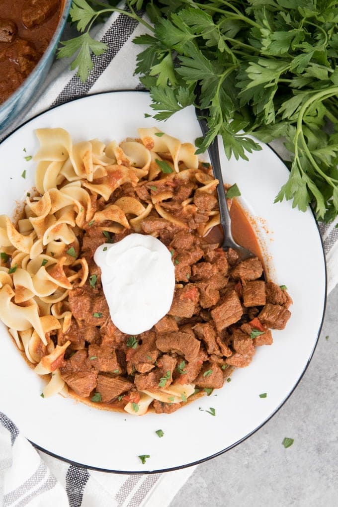 Authentic Hungarian Goulash with egg noodles and sour cream in a shallow white bowl with a black spoon.