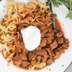 a square image of a white bowl with hungarian beef stew, egg noodles and sour cream