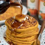 stack of pumpking pancakes on a white plate with butter and syrup pouring from a bottle.