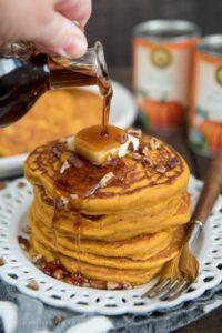 stack of pumpking pancakes on a white plate with butter and syrup pouring from a bottle.