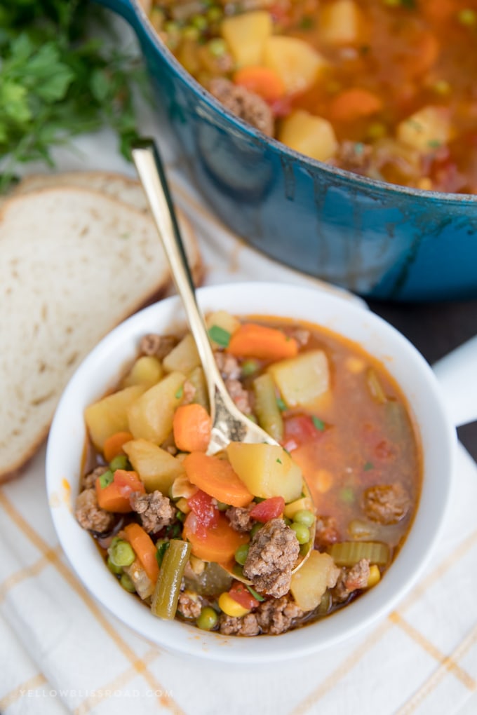 A bowl of Smoky Vegetable Beef Soup with a gold spoon, bread and a large blue pot.