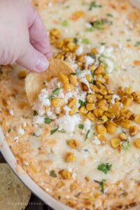mexican street corn dip, a hand holding a tortilla chip, roasted corn, cotija cheese and cilantro on top. image for social media