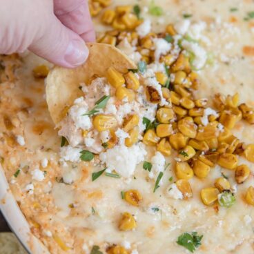 mexican street corn dip, a hand holding a tortilla chip, roasted corn, cotija cheese and cilantro on top. image for social media