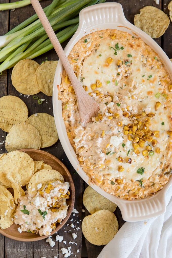 Hot Mexican Corn Dip has all the flavors of Mexican Street Corn in a deliciously dippable format with creamy cheeses and pops of sweet, roasted corn. Ceramic baking dish, cheesy mexican corn dip, green onions, wooden spoon, tortilla chips, wood plate