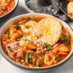 A bowl of soup with tomato broth, tortellini, spinach, cheese and parsley. A small piece on bread on top. Image created for social media.