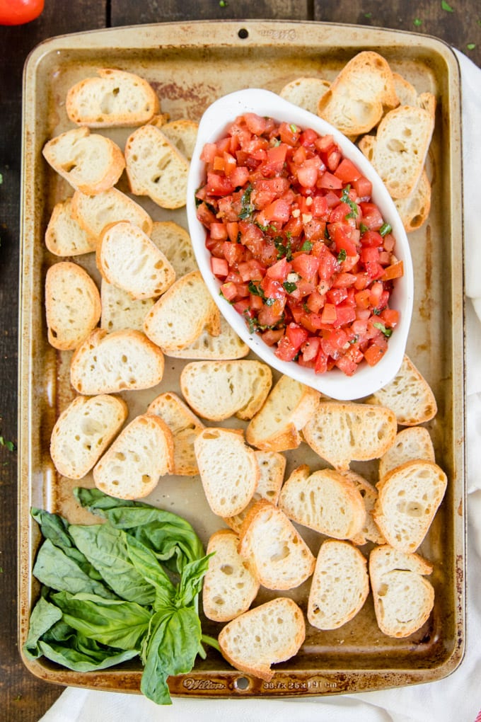 An overhead shot of a bowl of tomato bruschetta surrounded by fresh basil and toasted bread pieces.
