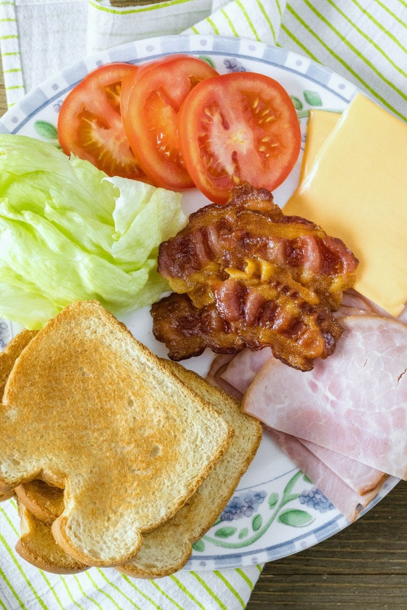 A stacked Club Sandwich is always a favorite at lunchtime. With two types of deli meat, cheese, lettuce, tomato, and bacon, what's not to love?! ingredients to make a club sandwich spread out on a plate