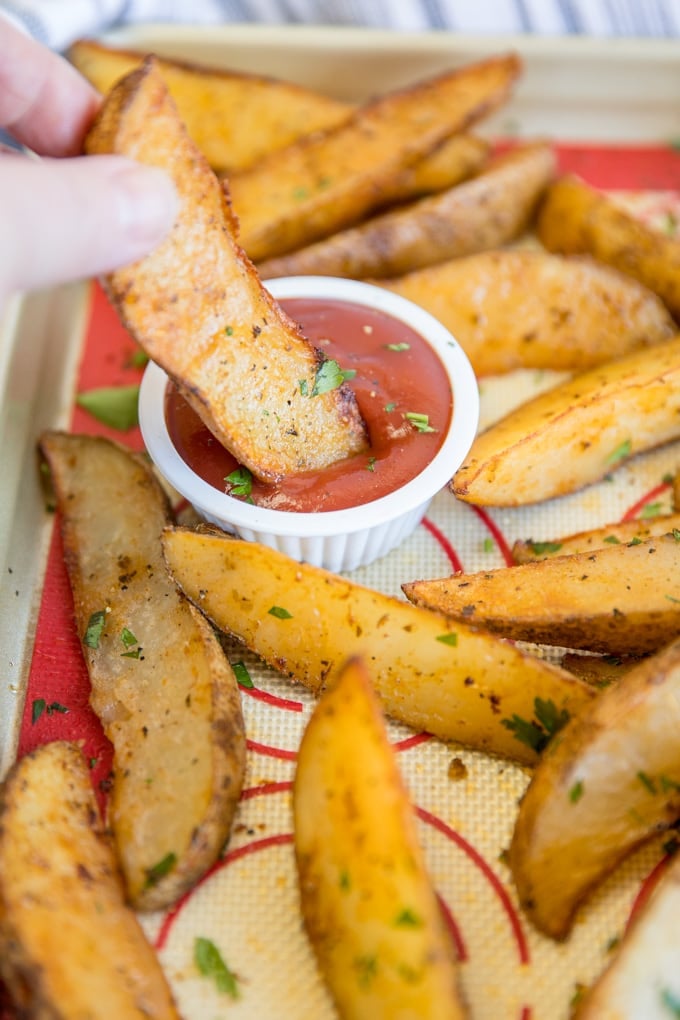 Baked potato wedges, with a hand dipping one into ketchup.