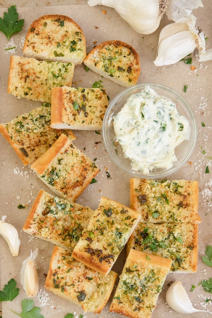 Sliced up garlic bread on parchment paper, surrounded by cloves of garlic and parsley