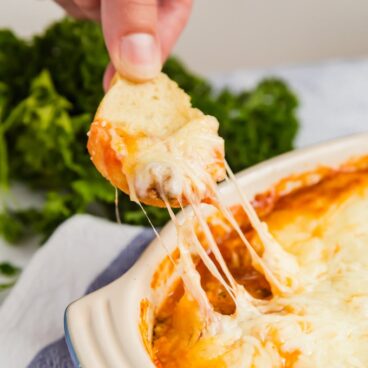 A hand pulling a sliced baguette with melted cheese from a dish of Lasagna Dip