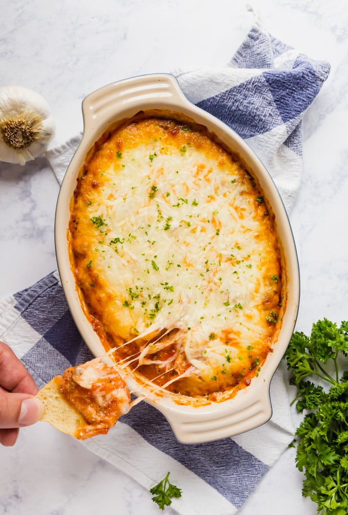 An overhead image of a dish of Lasagna dip with a hand taking some on a slice of baguette with melted cheese.