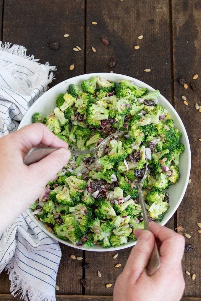 Two hands holding salad spoons to toss a broccoli salad.