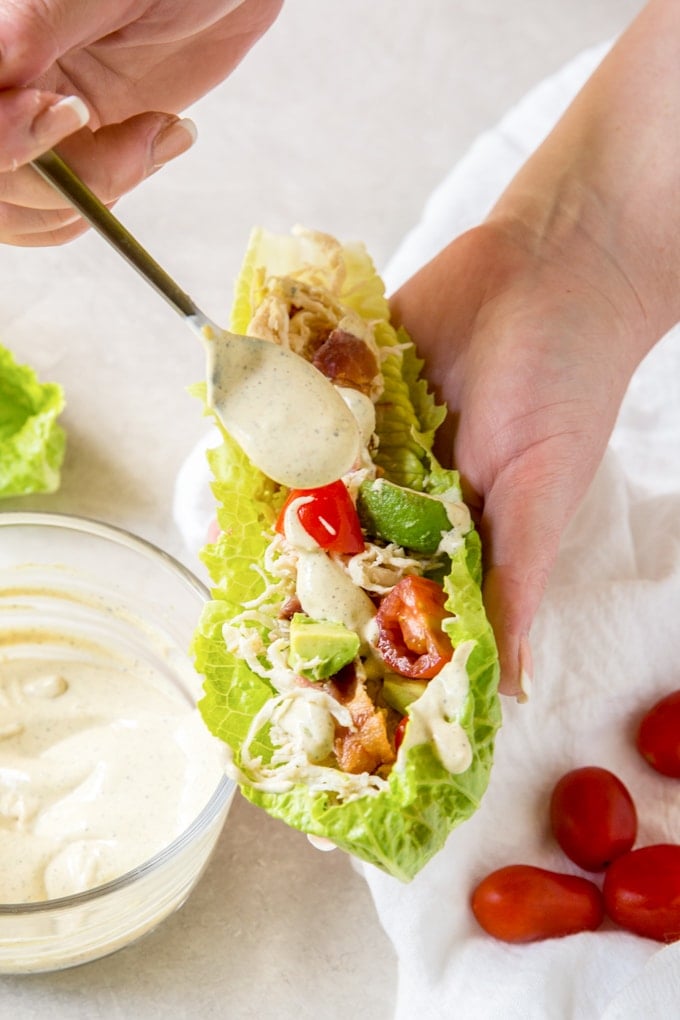 Tangy, crunchy, savory and light - these crunchy Chicken Club Lettuce Wraps are a perfect light lunch or even appetizer and they're good for you too! A hand holding a lettuce wrap and drizzling dressing on top