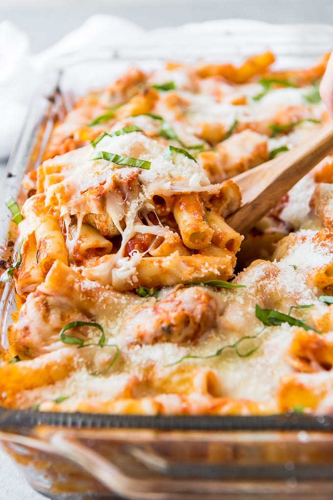 A close up of a wooden spoon lifting a serving of baked ziti out of a casserole dish