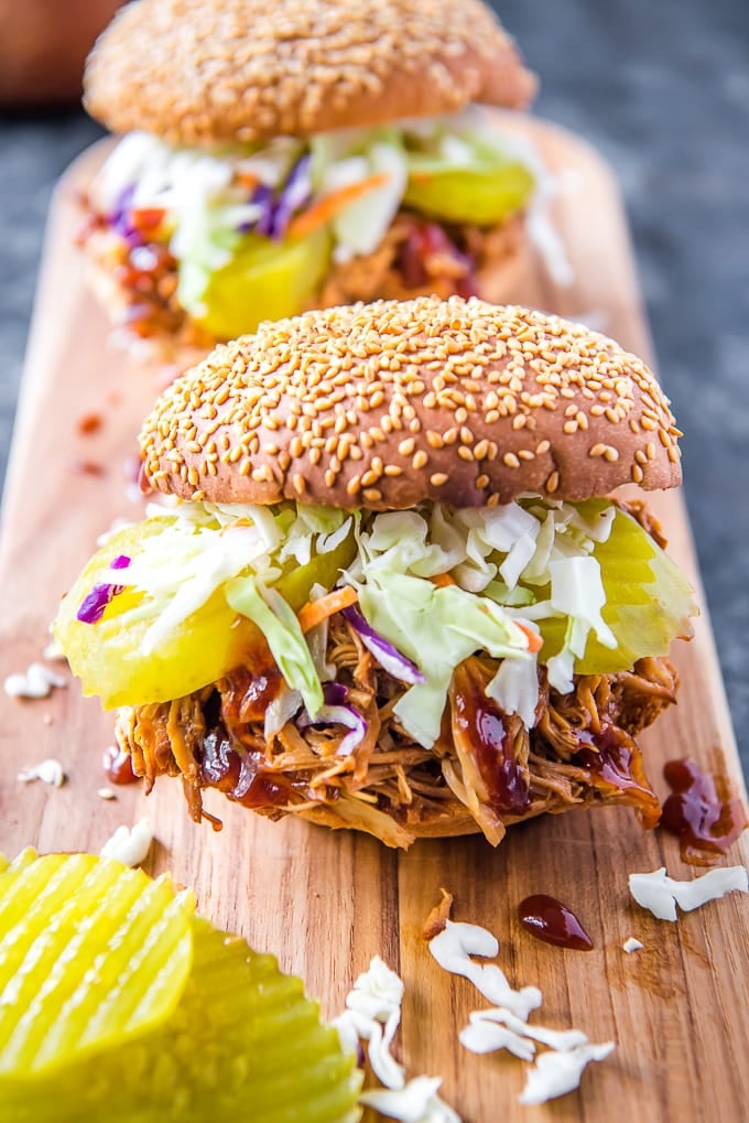 2 Crockpot BBQ Chicken piled onto sesame seed buns with coleslaw and pickles, sitting on a wooden cutting board.