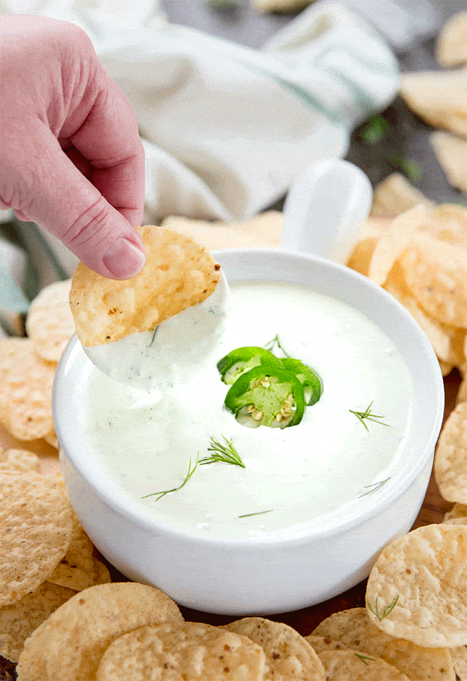A hand holding a tortilla chip with jalapeno dip dip in a bowl