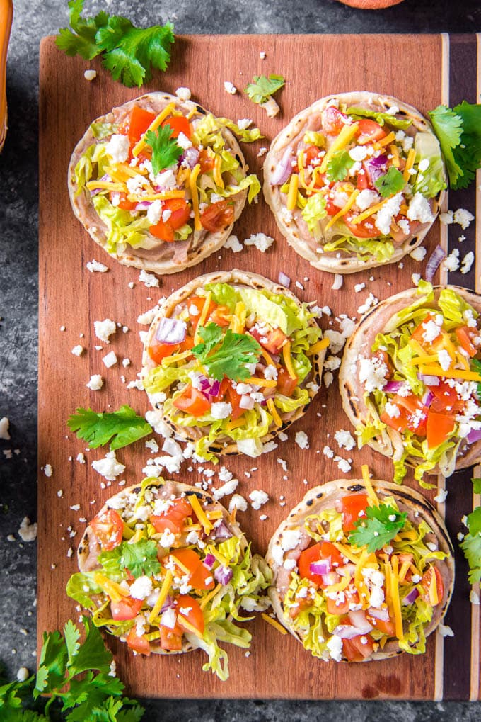 Authentic Sopes are a traditional Mexican dish that's perfect for Cinco de Mayo! Top them with beans, meat and vegetables for a delicious snack or meal. An overhead shot of Mexican sopes with toppings on a cutting board