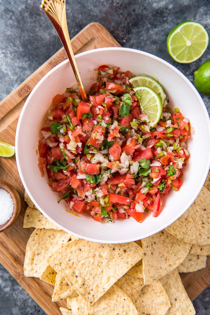 a white bowl filled with pico de gallo sitting on a cutting board surrounded by tortilla chips and limes.