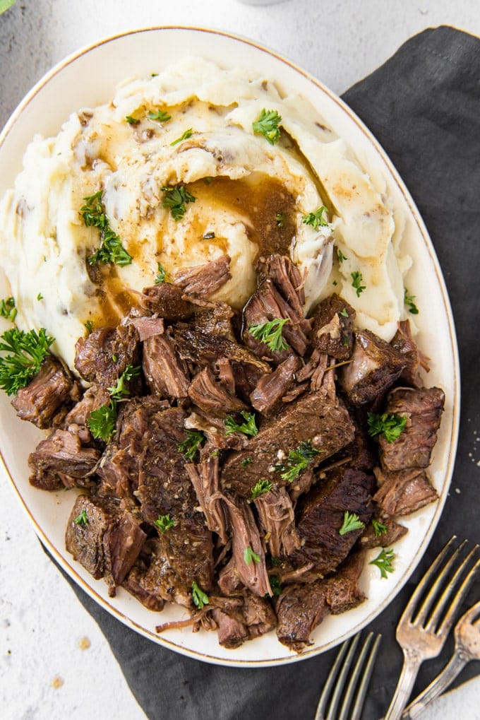 A platter of beef short ribs, slightly shredded, sitting next to mashed potatoes and gravy and sprinkled with parsley.