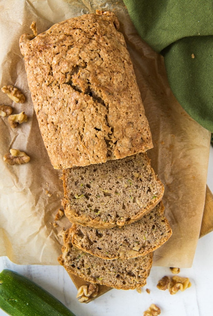 a loaf of zucchini bread, with three slices, parchment paper, walnuts, a zucchini and a green napkin.