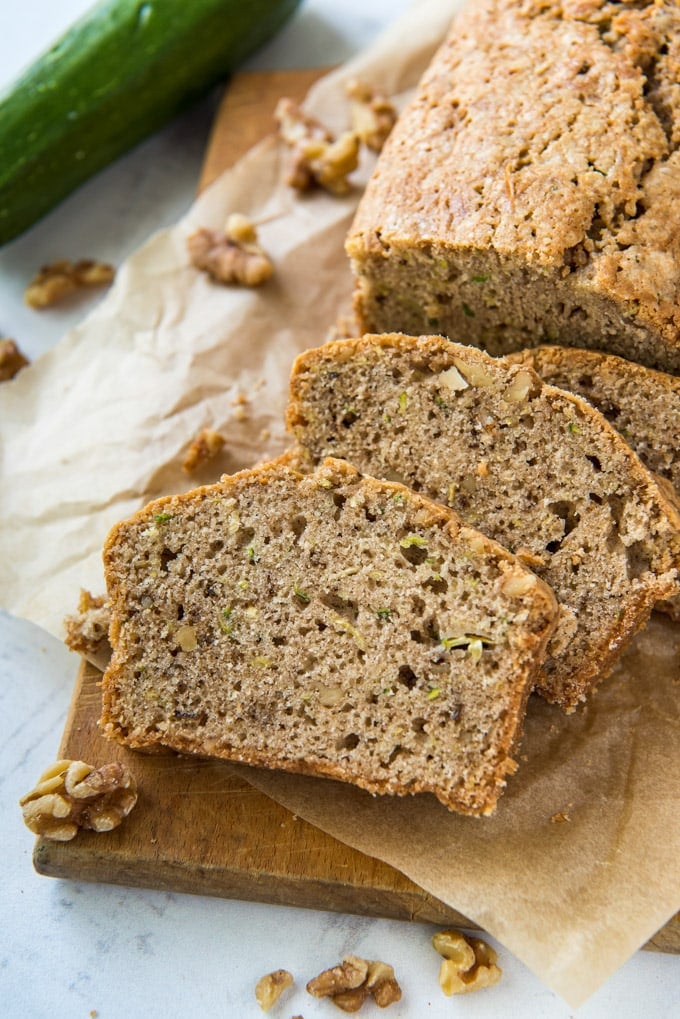 a loaf of zucchini bread, sliced, a zucchini, parchment paper, walnuts and a cutting board.