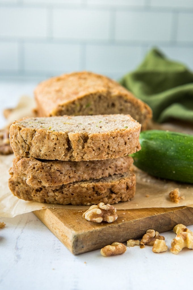 3 slices of zucchini bread stacked, a zucchini, a green napkin, a wood cutting board.