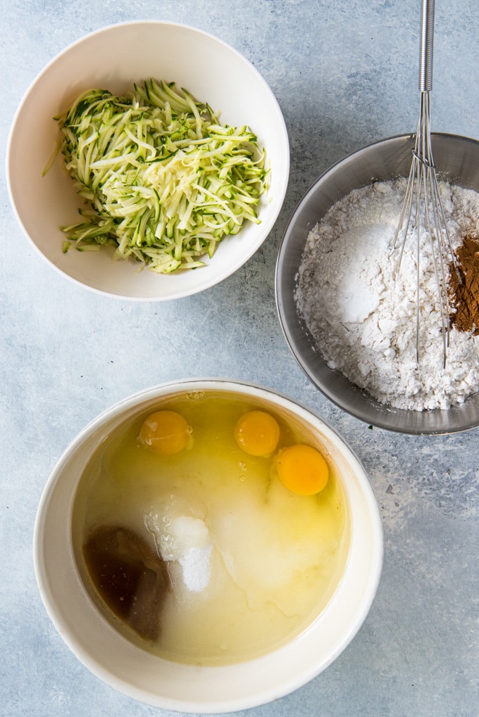 3 bowls with shredded zucchini, flour & cinnamon, and a bowl with eggs