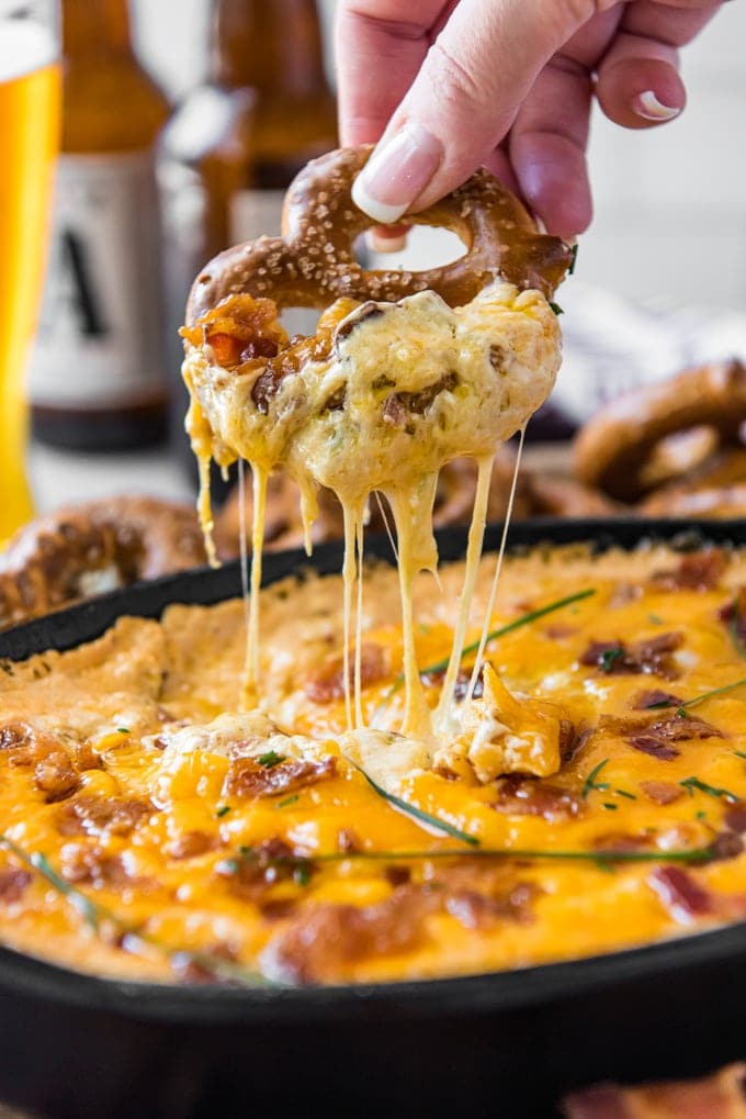 A large hard pretzels dripping with stringy cheese from a skillet of bacon beer cheese dip.