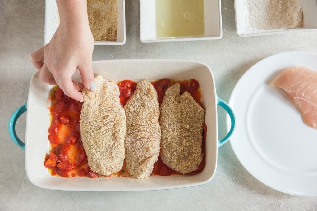 A hand placing a breaded chicken breast in a baking pan with a thin layer of marinara.