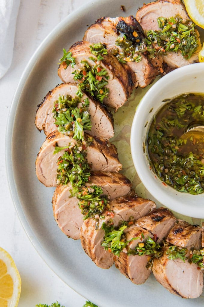 A platter of sliced grilled pork tenderloin drizzled with chimichurri sauce.