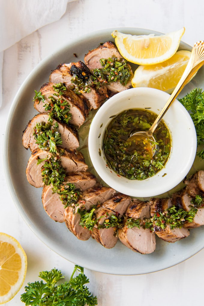 An overhead shot of slices of grilled pork tenderloin surrounded a dish of chimichurri sauce