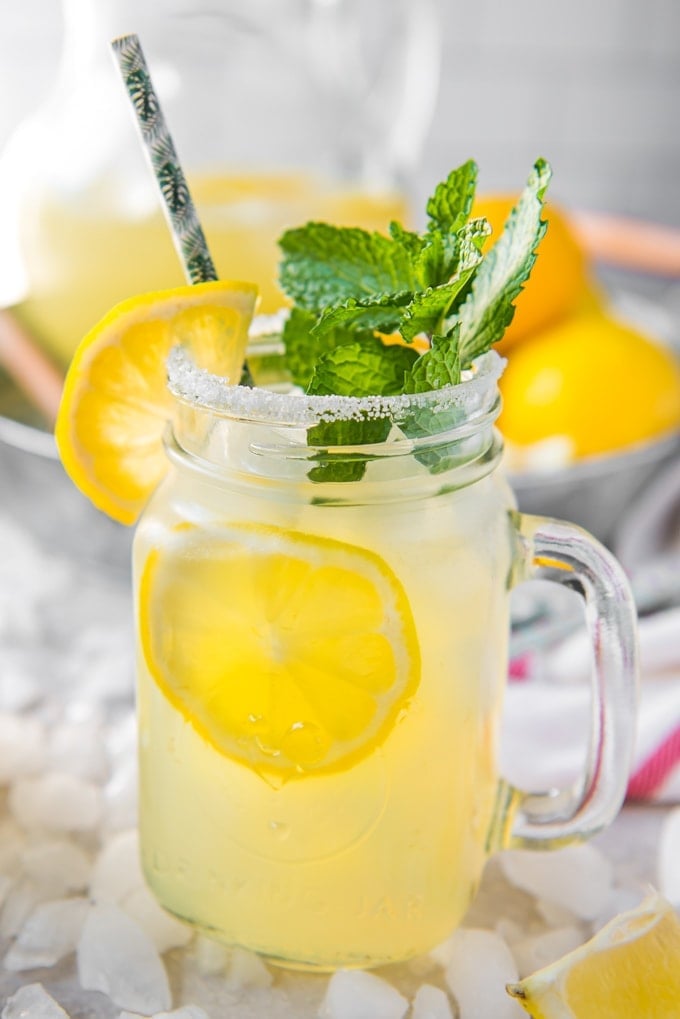 A close up of a pretty glass mug with lemonade , lemon slices, mint leaves and a straw.