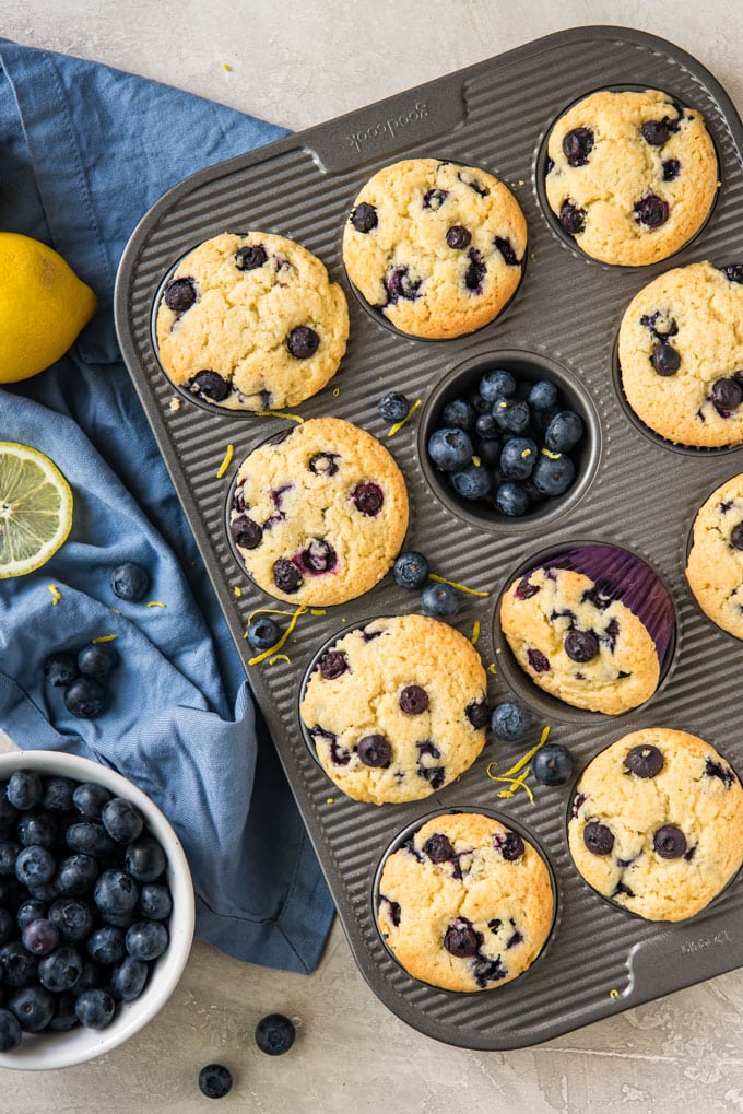 An overhead shot of a muffin pan full of lemon blueberry muffins, a bowl of blueberries and a couple of lemons on a blue towel.